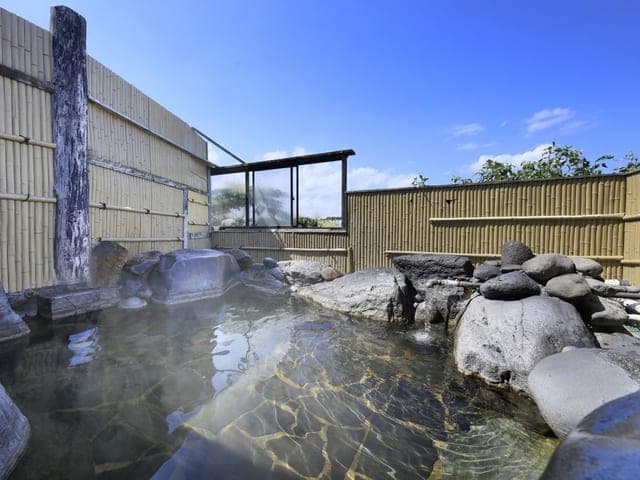 岩を組んだ露天風呂。湯気の向こうに木の塀と青空が見える