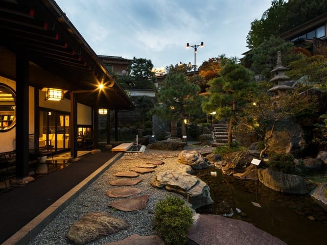 夕暮れの日本庭園。灯籠と軒の灯りが、石組みと植栽の間に点々とともる