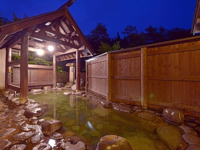 夕暮れの露天風呂「鶴の湯」。木塀に囲まれた湯船に灯りがともる